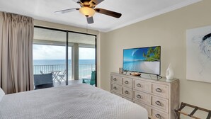 A hotel room with a large bed, a ceiling fan, a TV on a dresser, a painting on the wall, and a view of the ocean through the balcony doors. - White Caps 1202 by Liquid Life (Orange Beach)