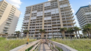 A multi-story hotel with a central courtyard, balconies, and a wooden walkway leading to the entrance. - White Caps 1202 by Liquid Life (Orange Beach)