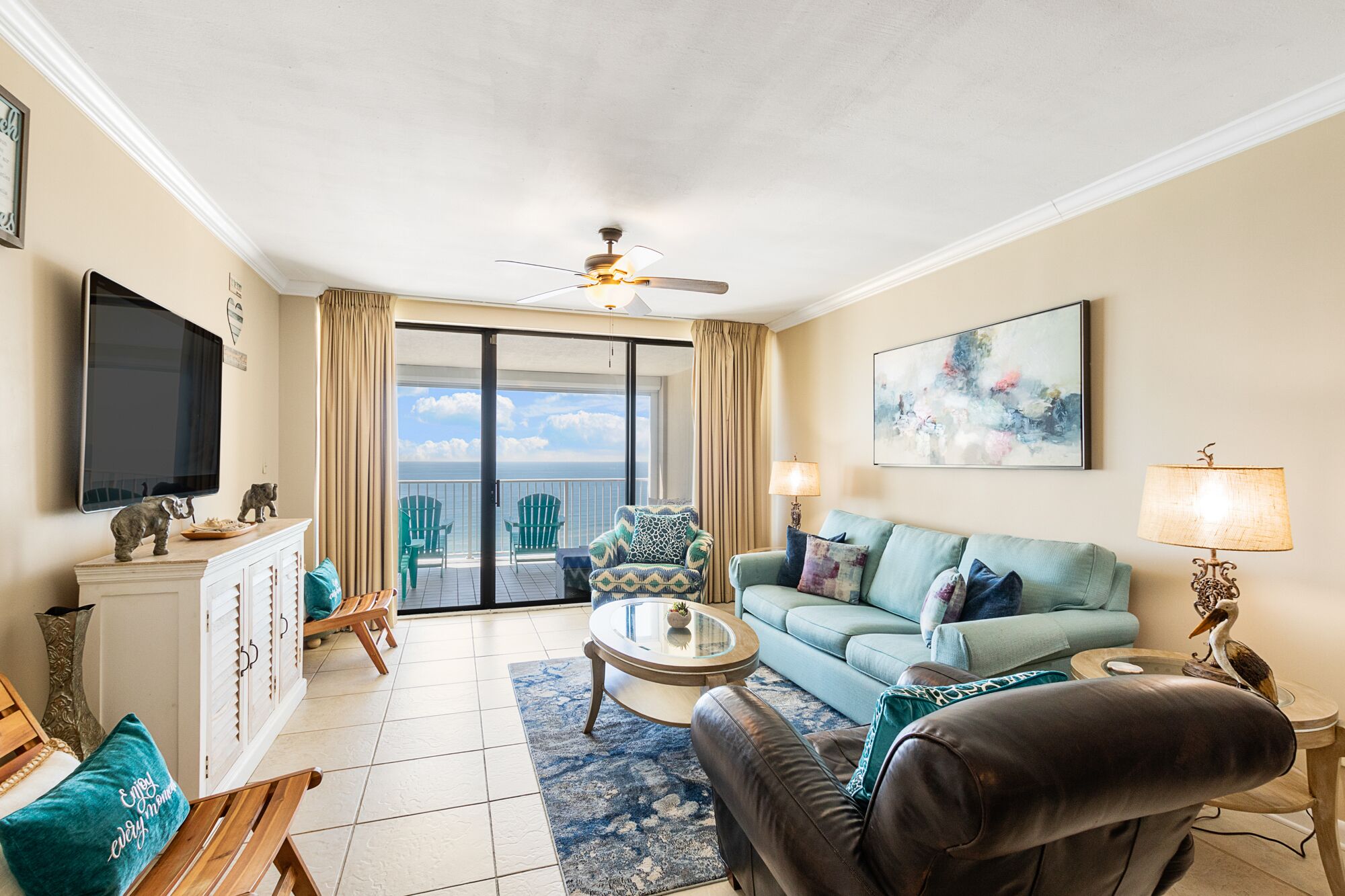 Ein Wohnzimmer mit einer Couch, Sesseln, einem Couchtisch und einem Fernseher. Es gibt einen Balkon mit Stühlen und Blick aufs Meer.