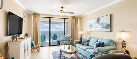 Ein Wohnzimmer mit einer Couch, Sesseln, einem Couchtisch und einem Fernseher. Es gibt einen Balkon mit Stühlen und Blick aufs Meer.