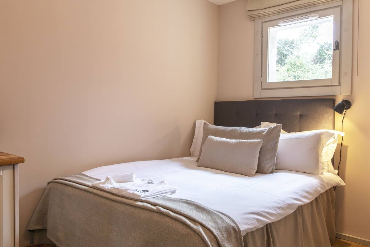 A neatly made bed with a headboard, a bedside lamp, and a window with a view of greenery.