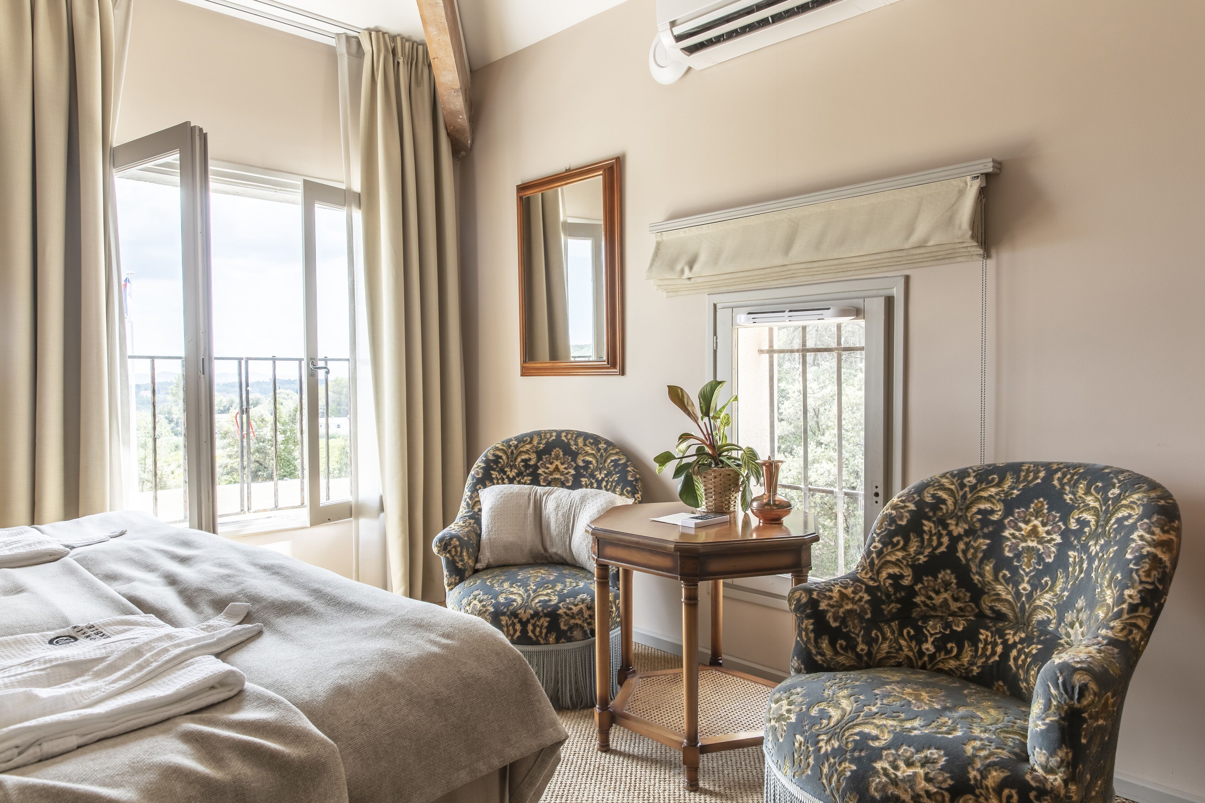 A hotel room with a bed, two armchairs, a small table with a plant, and a window with curtains.
