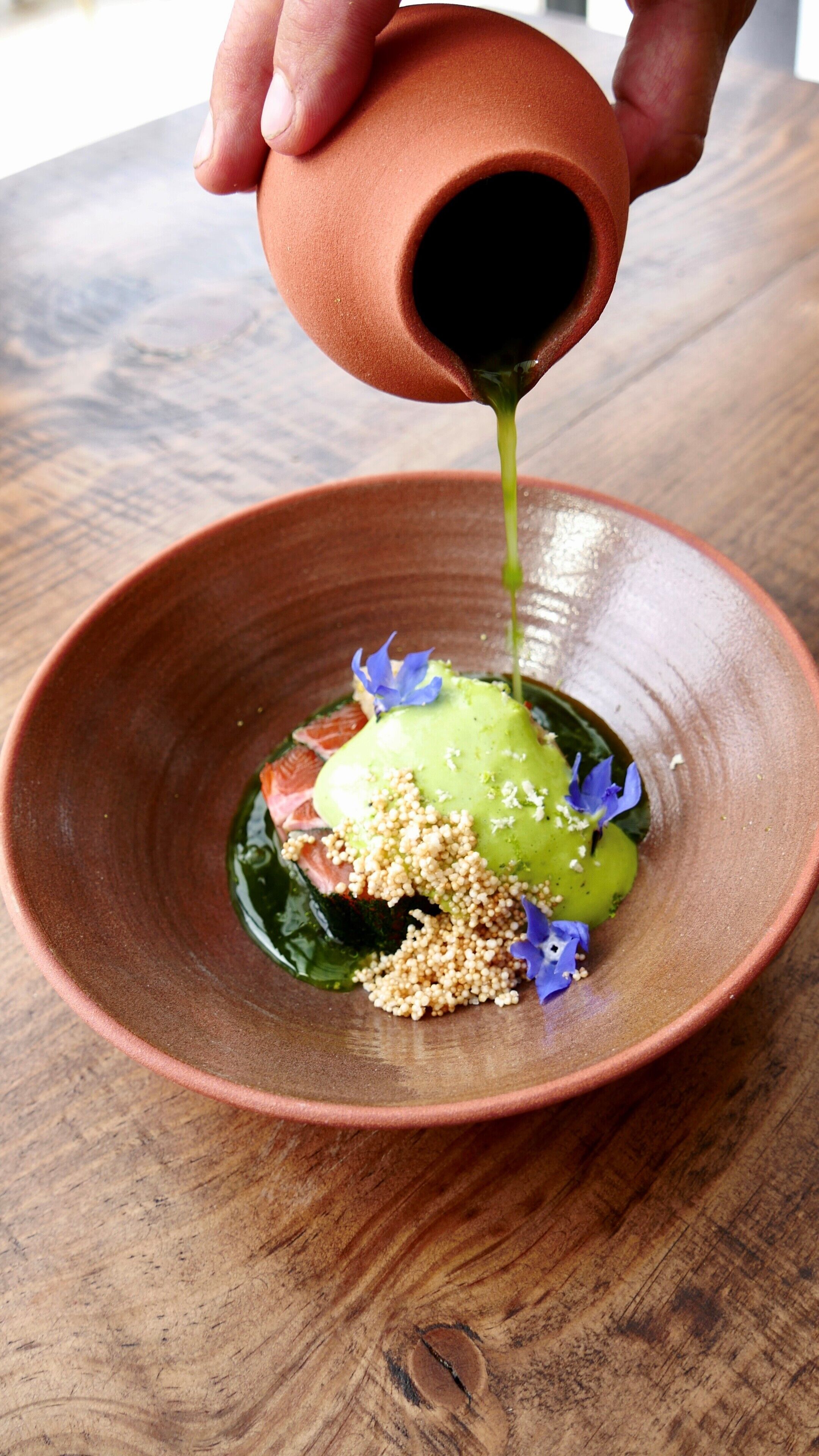 A hand pouring a green sauce into a bowl containing a dish with various ingredients.