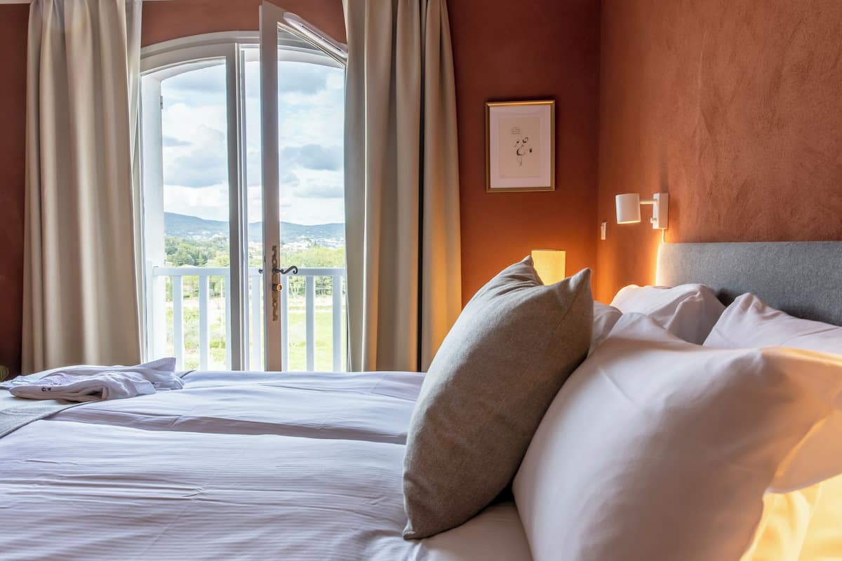 A hotel room with a bed, a view of the outdoors, and a framed picture on the wall.