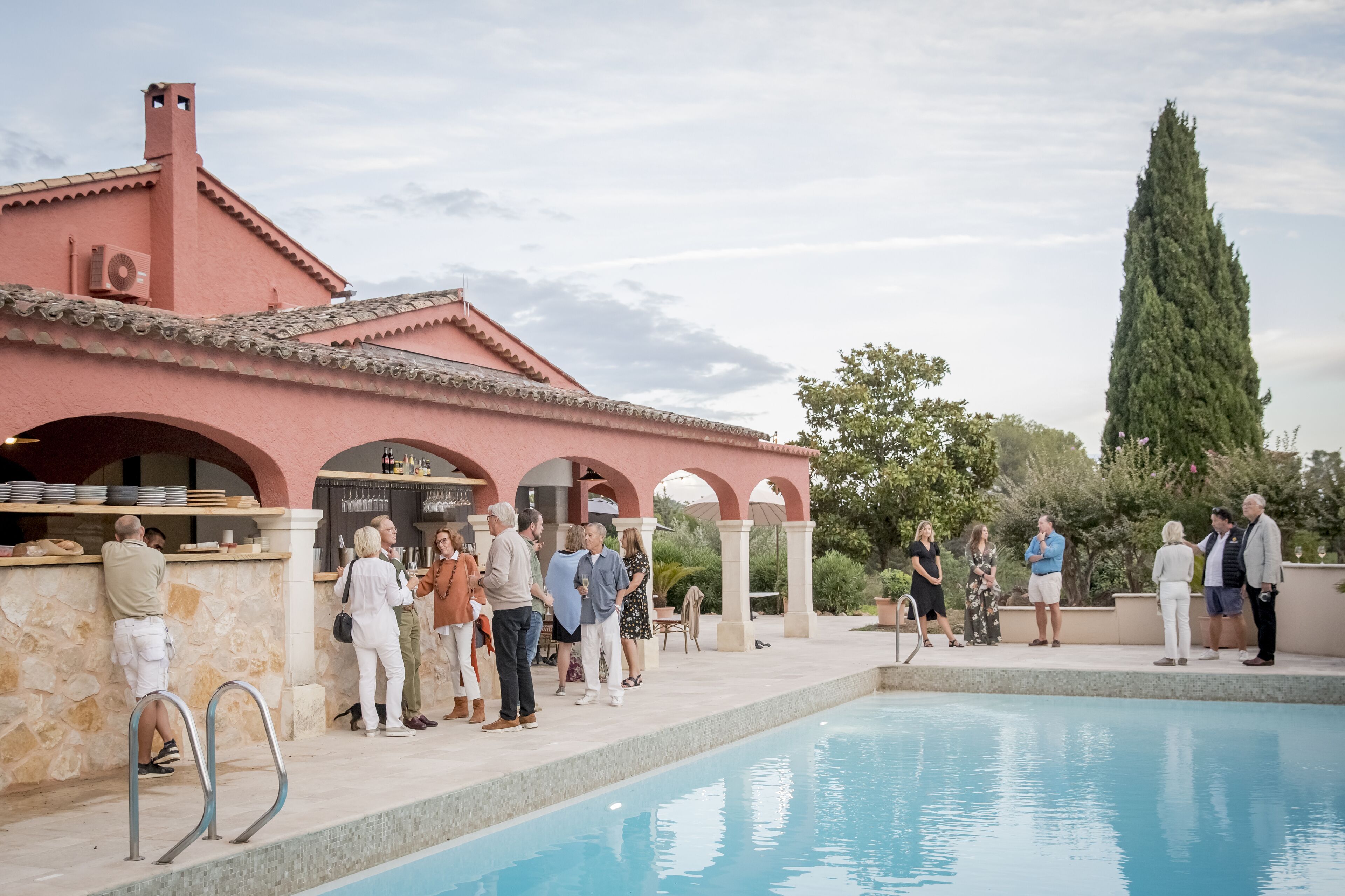 Un espace aménagé au bord de la piscine, avec un bâtiment en pierre, un grand arbre et un ciel dégagé.