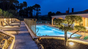 Une piscine avec des chaises longues et un petit arbre, le tout entouré d’un mur de pierre et d’un bâtiment doté d’un toit en tuiles.