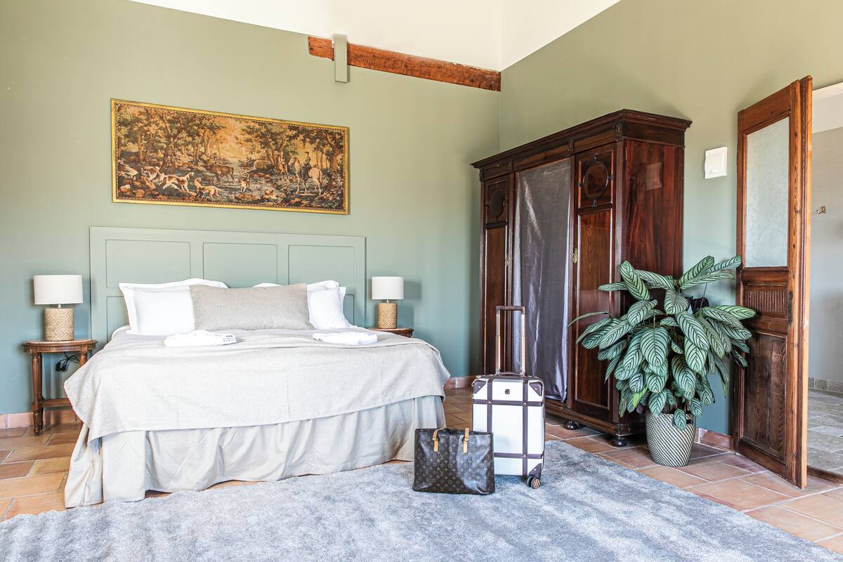 A bedroom with a bed, bedside tables, a large painting, a wardrobe, a suitcase, and a potted plant.
