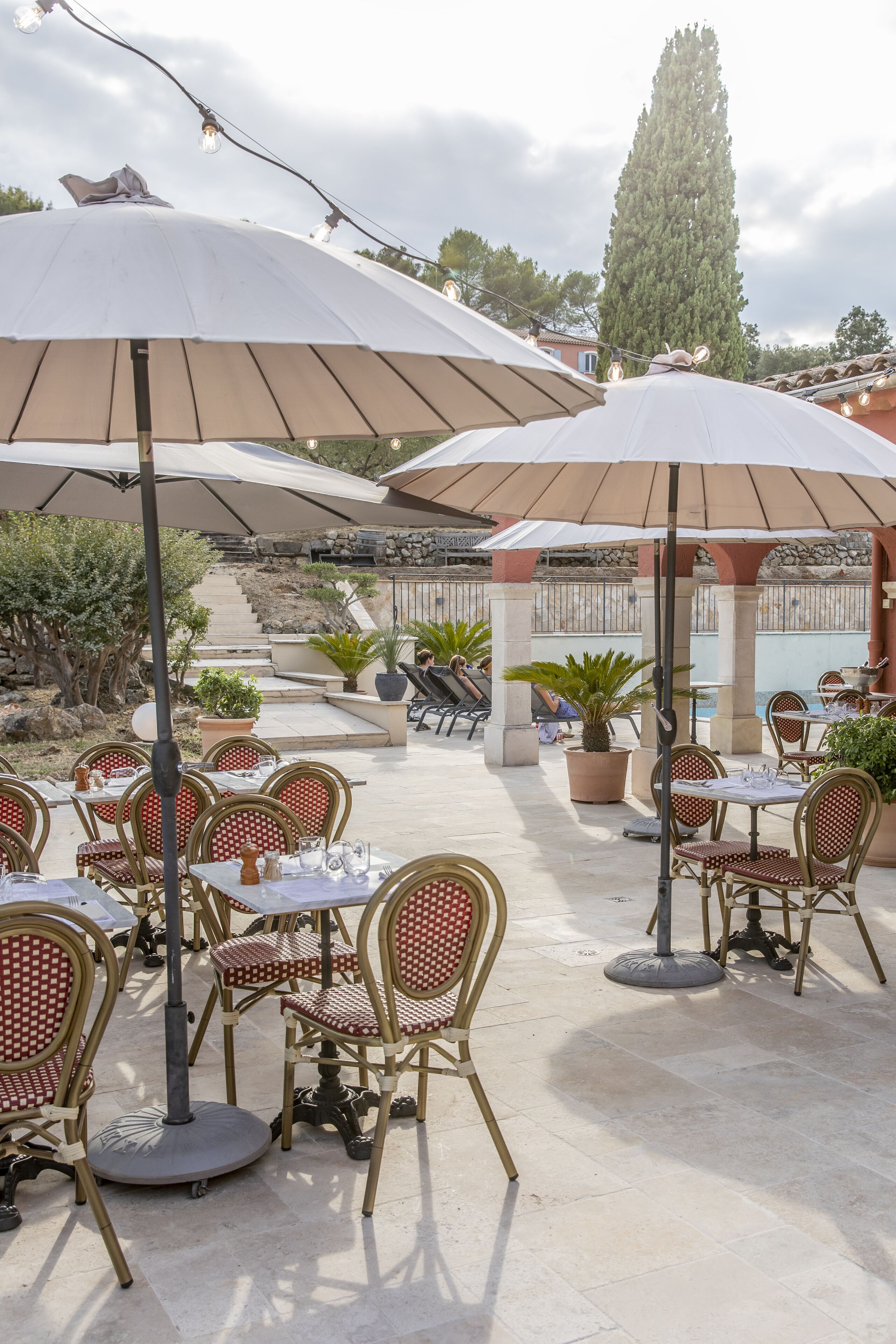 Een terras buiten met meerdere tafels en stoelen, waarvan sommigen onder parasols staan, en potplanten eromheen.