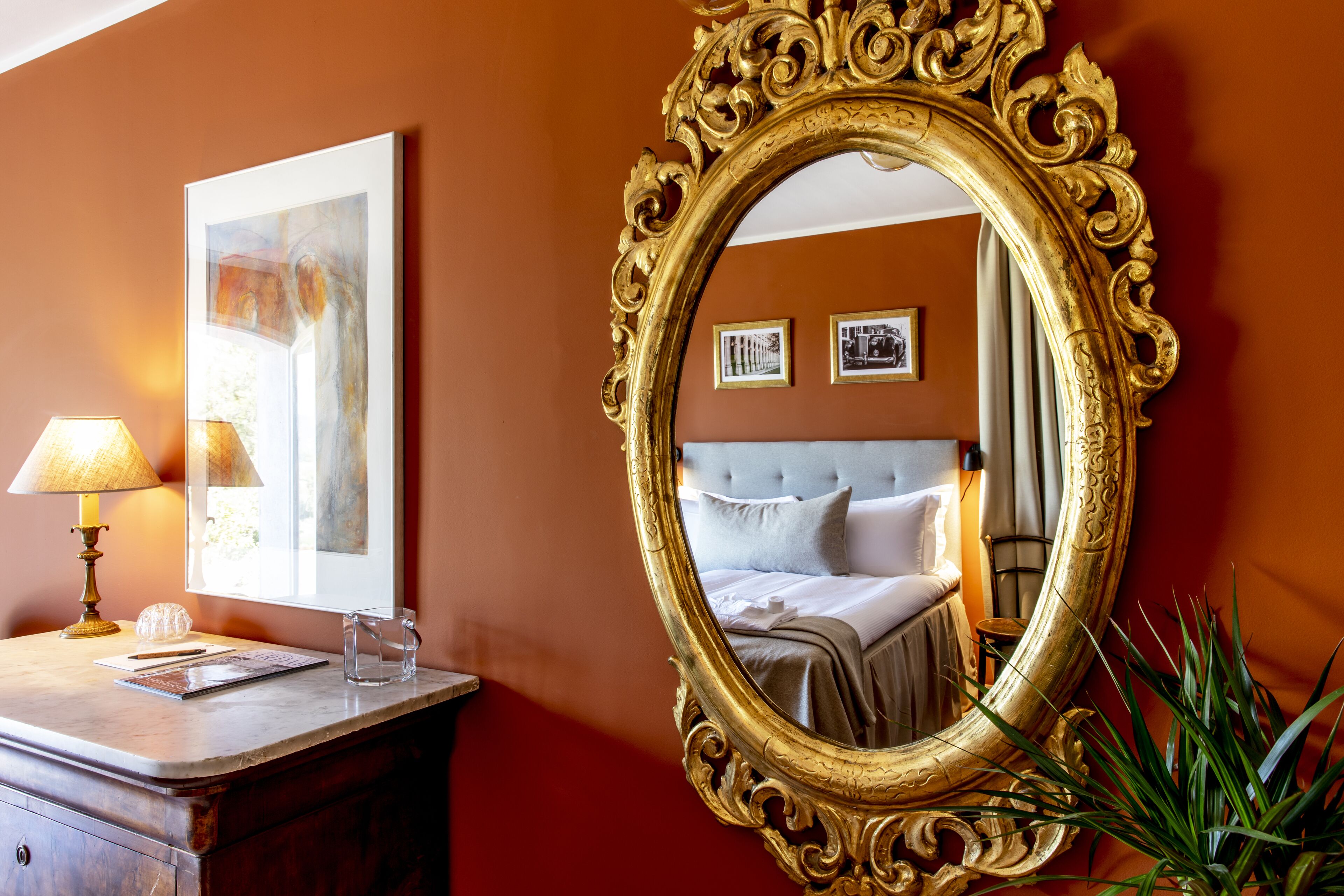 Habitación de hotel con un espejo grande y decorativo, una mesita de noche con lámpara, un cuadro enmarco y una cama con cabecero gris.