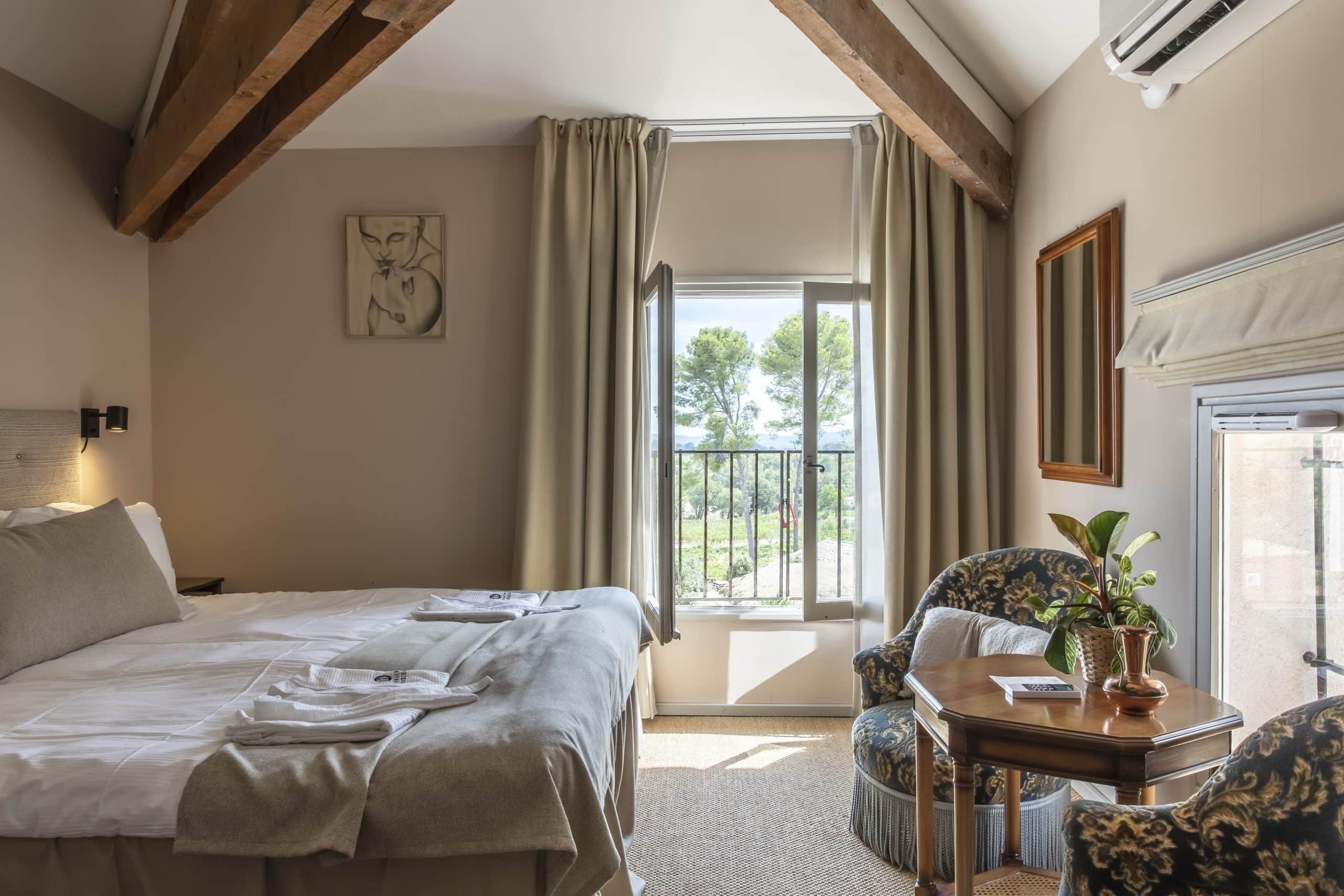 A hotel room with a large bed, a chair, a small table with a plant, and a balcony with curtains.