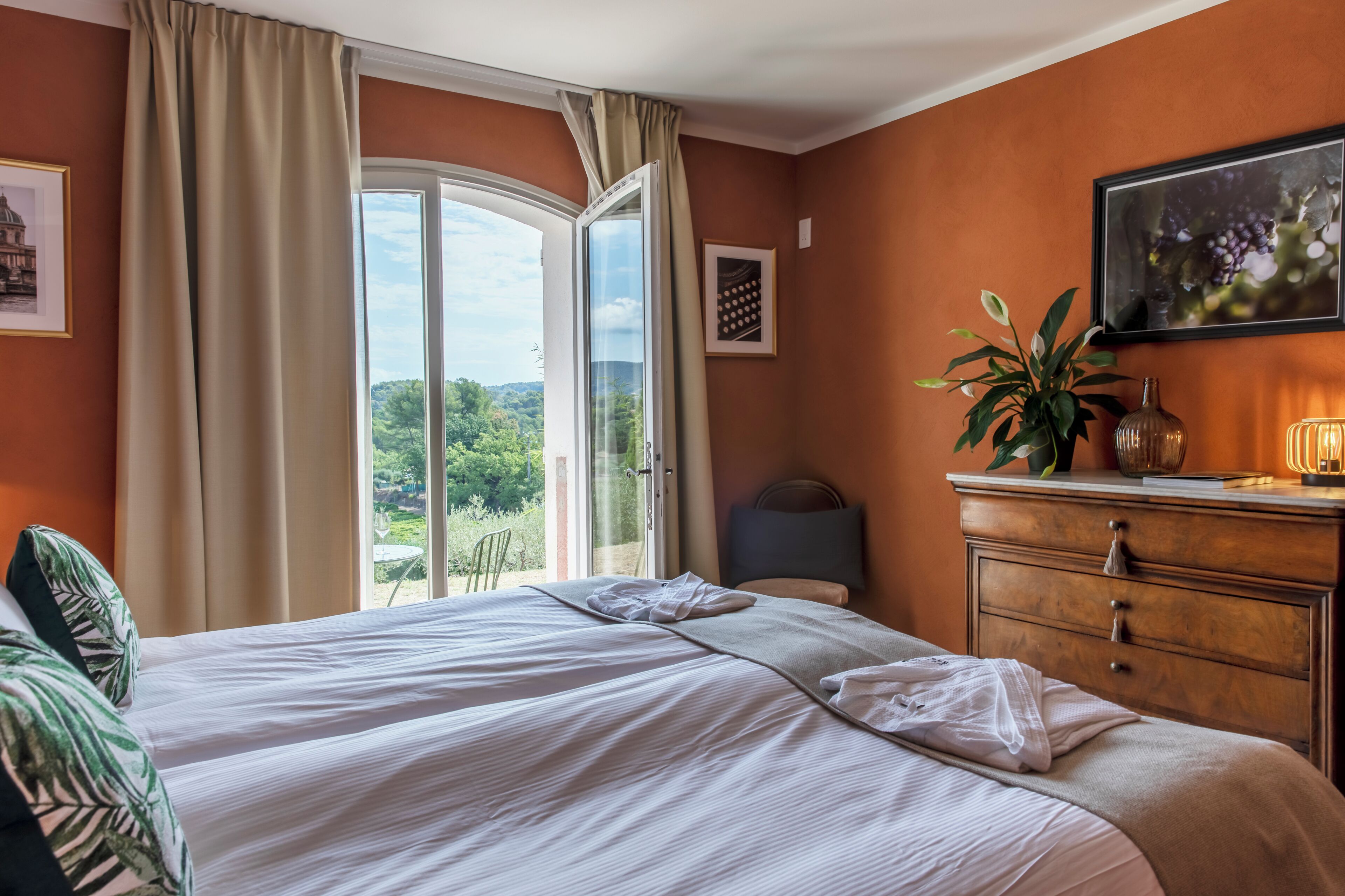 A hotel room with a bed, a dresser, a plant, and a view of greenery outside.
