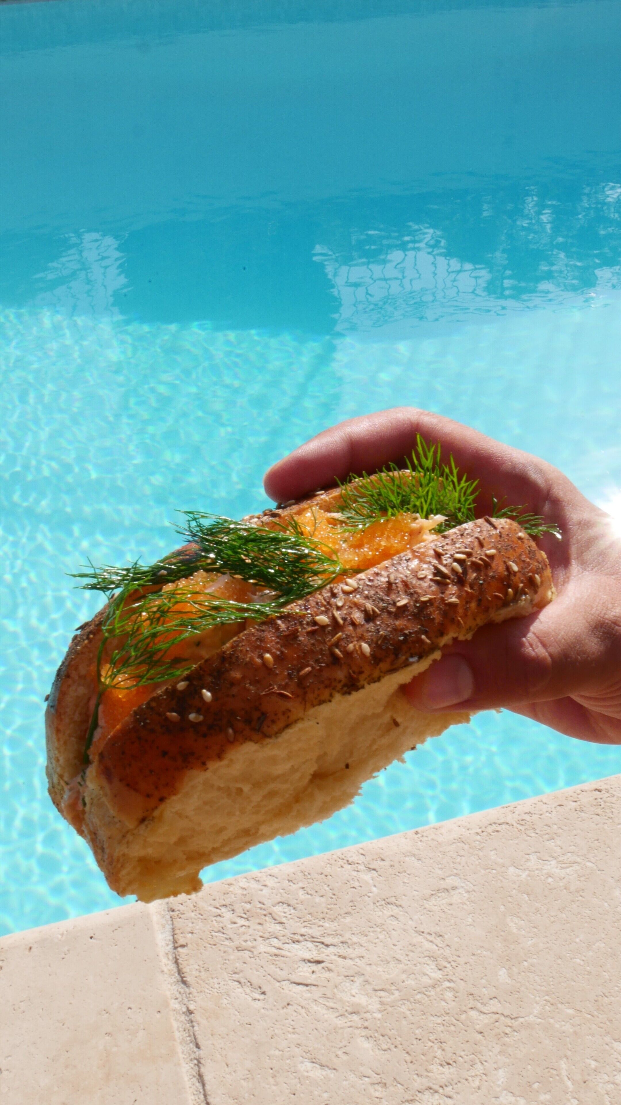 Une main tenant une tranche de pain garnie de saumon et d’aneth, près d’une piscine.