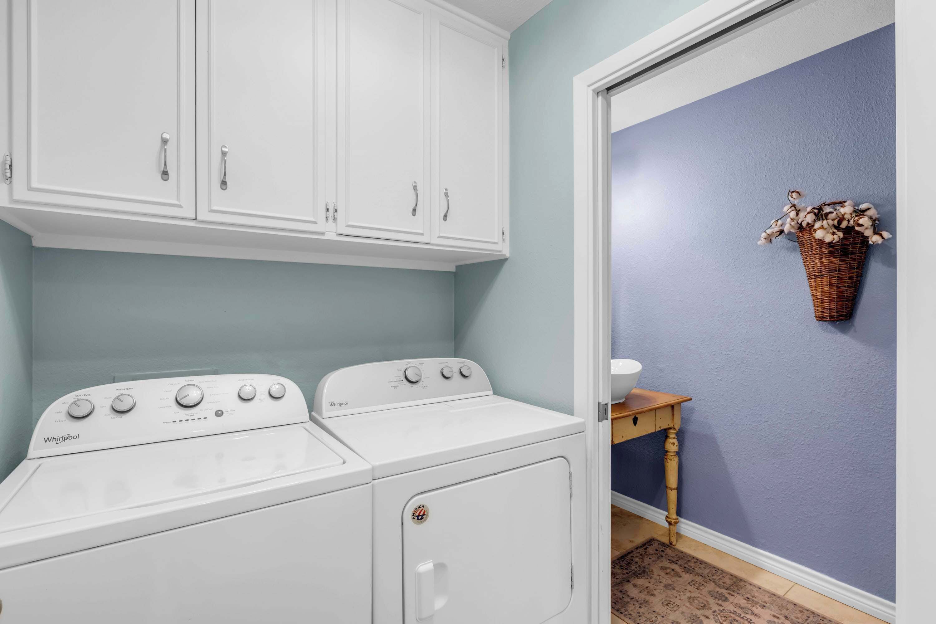 Een wasruimte met een Whirlpool wasmachine en droger, een houten tafel met een mand vol bloemen, en een blauwe muur.