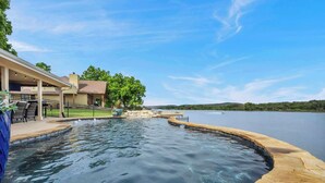 Ein Pool mit Steinrand, eine überdachte Terrasse mit Gartenmöbeln und Blick auf einen See und entfernte Hügel.