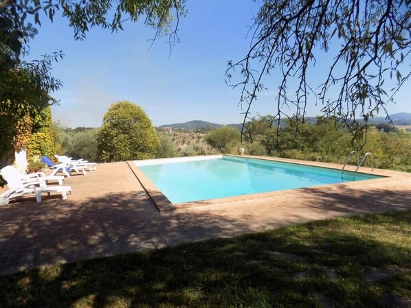 A rectangular swimming pool with a clear blue water surface, surrounded by a paved deck. There are several white lounge chairs with blue cushions arranged around the pool. The area is shaded by trees, and the background features a hilly landscape with greenery. - Scappo in Umbria, Il Fienile (Fornole)