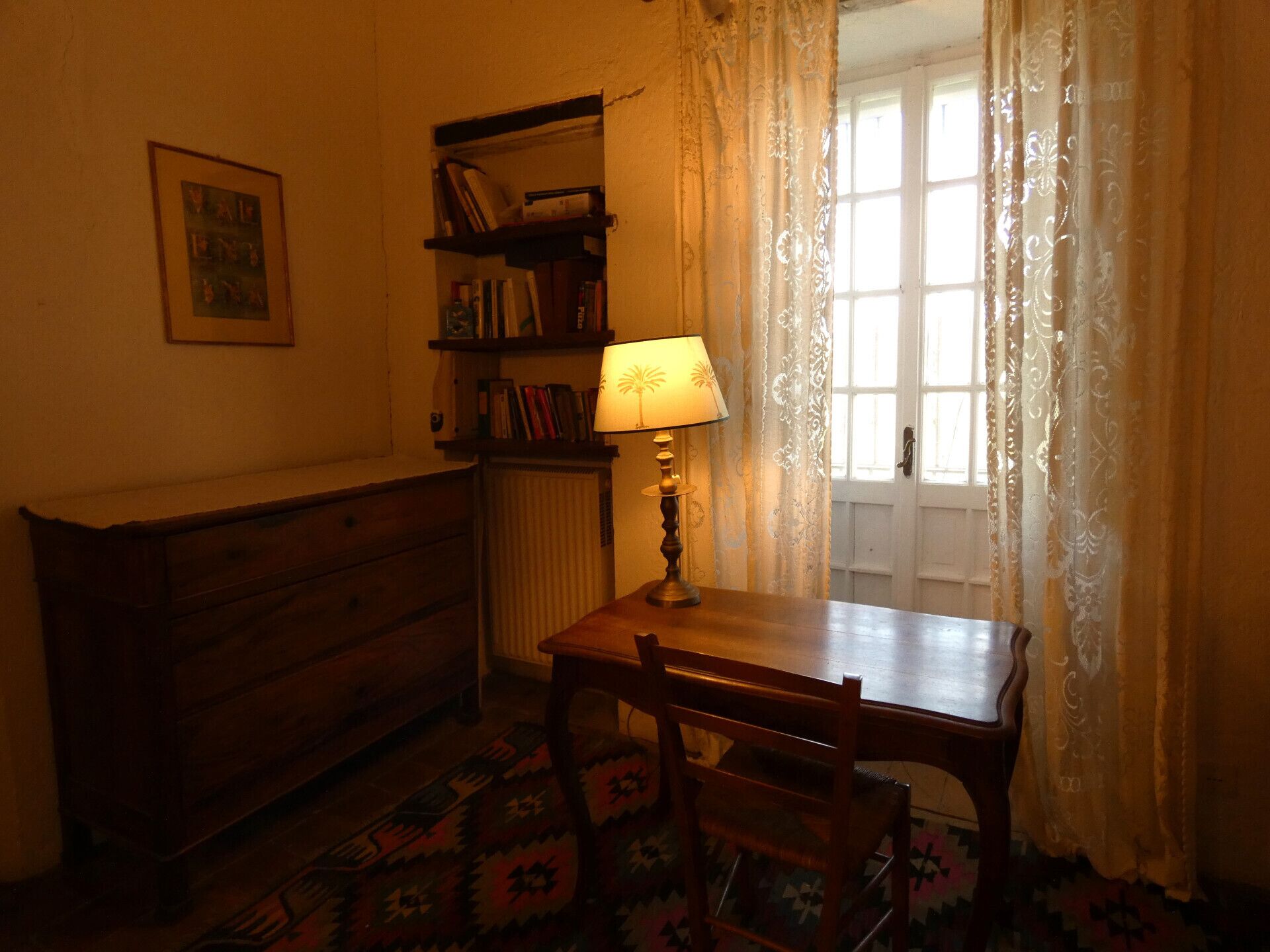 Una habitació amb un escriptori de fusta, una làmpada, una estanteria, un radiador i una catifa amb dibuixos.