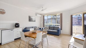 Une cuisine moderne avec des armoires blanches, un lave-vaisselle et une machine à café. On y trouve un coin repas avec une table en bois et des chaises blanches. Un canapé bleu avec des coussins à motifs est placé contre le mur, et un ventilateur de plafond est visible.