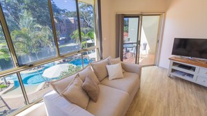 A living room with a large window, a sofa, a TV stand, and a view of a swimming pool and buildings. - Martys Little Beach No11 (Nelson Bay)