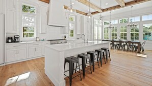 A spacious kitchen with white cabinetry, a large island with bar stools, wooden beams on the ceiling, and large windows providing natural light. - Private Neighborhood beach access, large pool, golf cart, walk to beach/dine (Santa Rosa Beach)