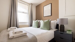 A neatly made bed with folded towels, a bedside lamp, and a small plant on a nightstand. - The Clerkenwell Apartments. (London)