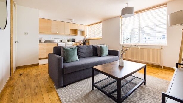 A modern apartment with a grey sofa, a wooden floor, a kitchen with wooden cabinets, and a large window with blinds. - The Clerkenwell Apartments. (London)
