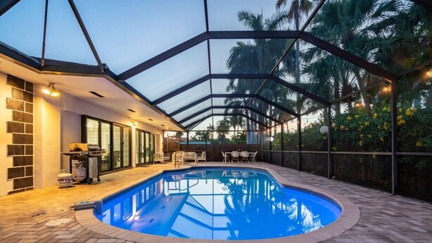 Una piscina techada con techo de vidrio, rodeada por un patio de ladrillo y mobiliario exterior.