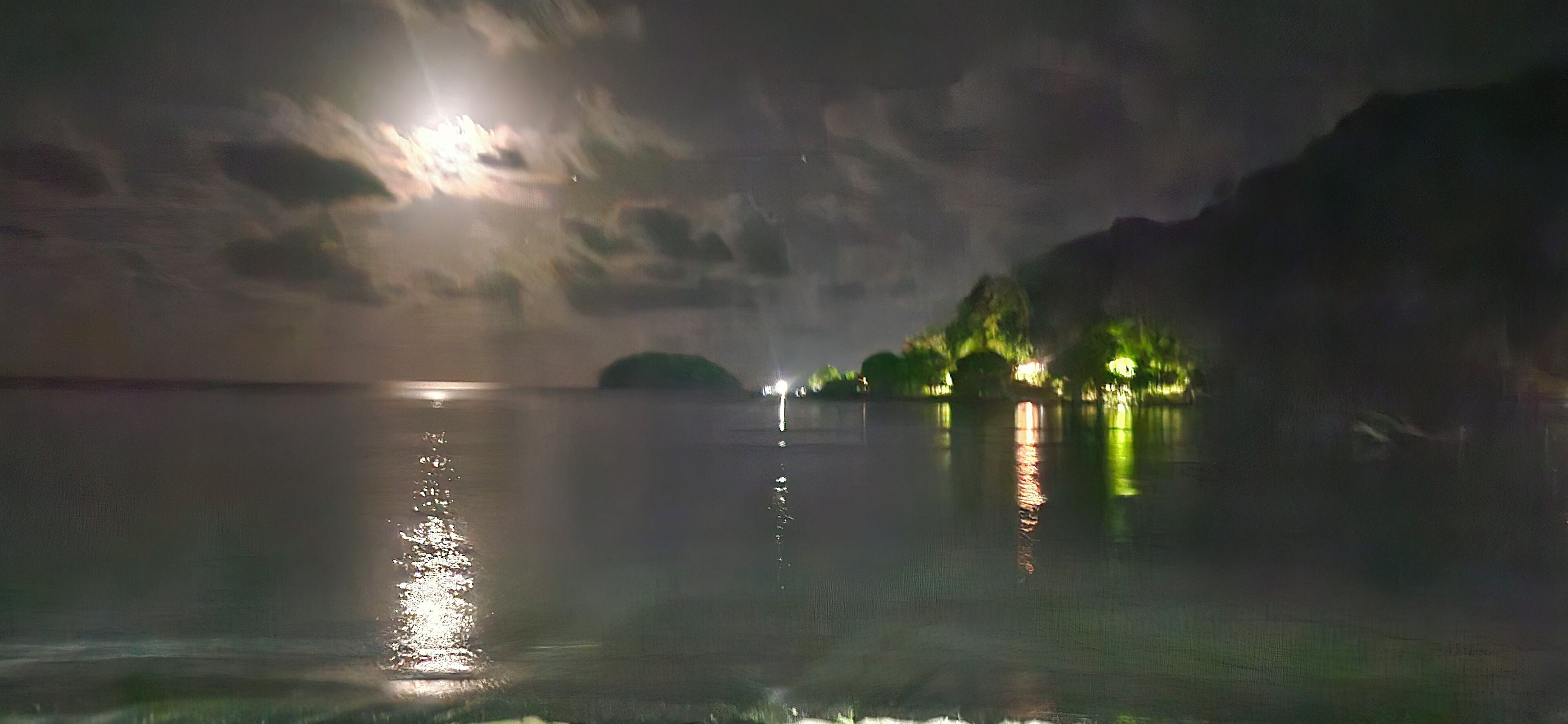 A nighttime view of a calm body of water reflecting a bright moon and distant lights.