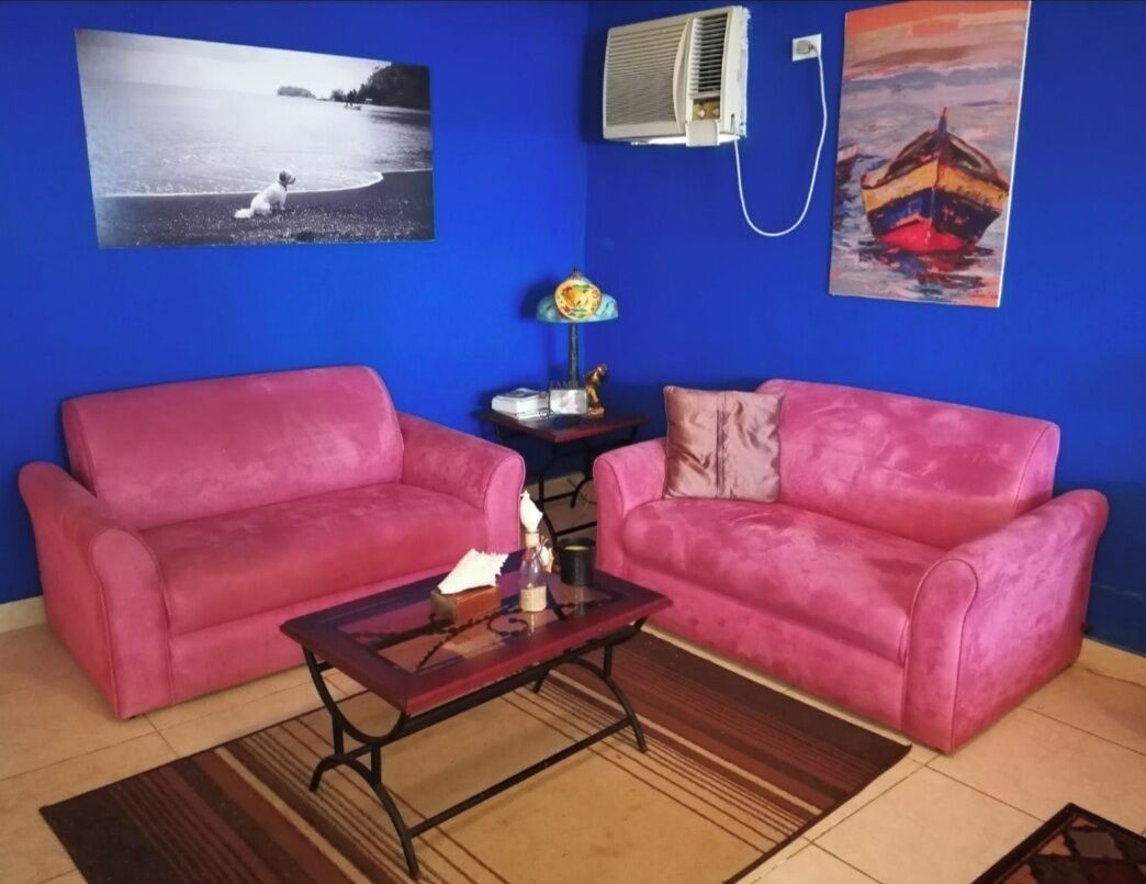 Ett rum med två rosa soffor, ett soffbord i trä och ett litet sidobord med en lampa. Det finns två väggmonterade luftkonditioneringsapparater och två inramade tavlor, en med en strandscen och en med en båt.