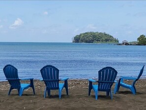 A beach with three blue chairs facing the sea, a small hut in the distance, and a lush green island. - BEAUTIFUL APARTMENT RIGHT BY THE OCEAN (Portobelo)
