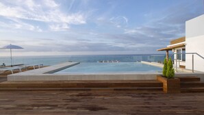 A rooftop infinity pool with a wooden deck, a small potted plant, and a glass railing overlooking the ocean.