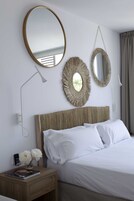A neatly made bed with white linens, a wooden nightstand with a book and flowers, and two wall-mounted mirrors with woven frames.