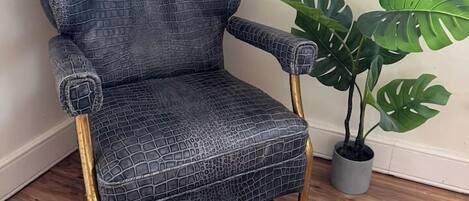 A corner chair with a textured fabric and gold legs, positioned next to a potted plant with broad leaves.