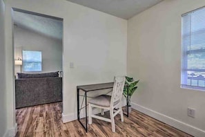 Una habitación con piso de madera, un escritorio blanco con una silla, un sofá gris y una ventana con persianas.