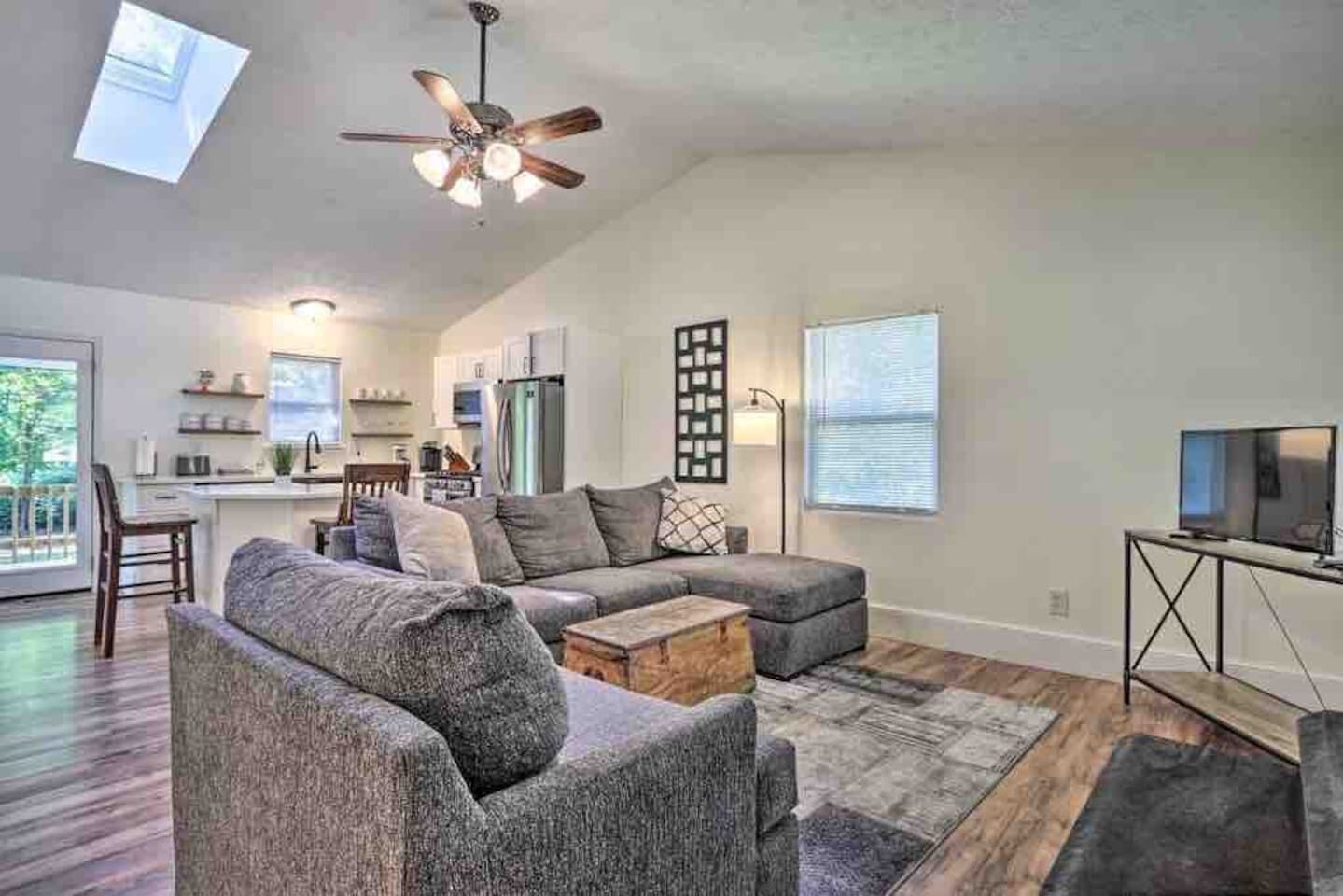 A spacious living room with a sectional sofa, a coffee table, and a TV on a stand. There is a ceiling fan and a kitchen area in the background.