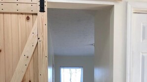 Ein enger Flur mit einer Holztür links, einem Bett mit weißer Bettwäsche in der Mitte und einem Fenster mit Tageslicht.