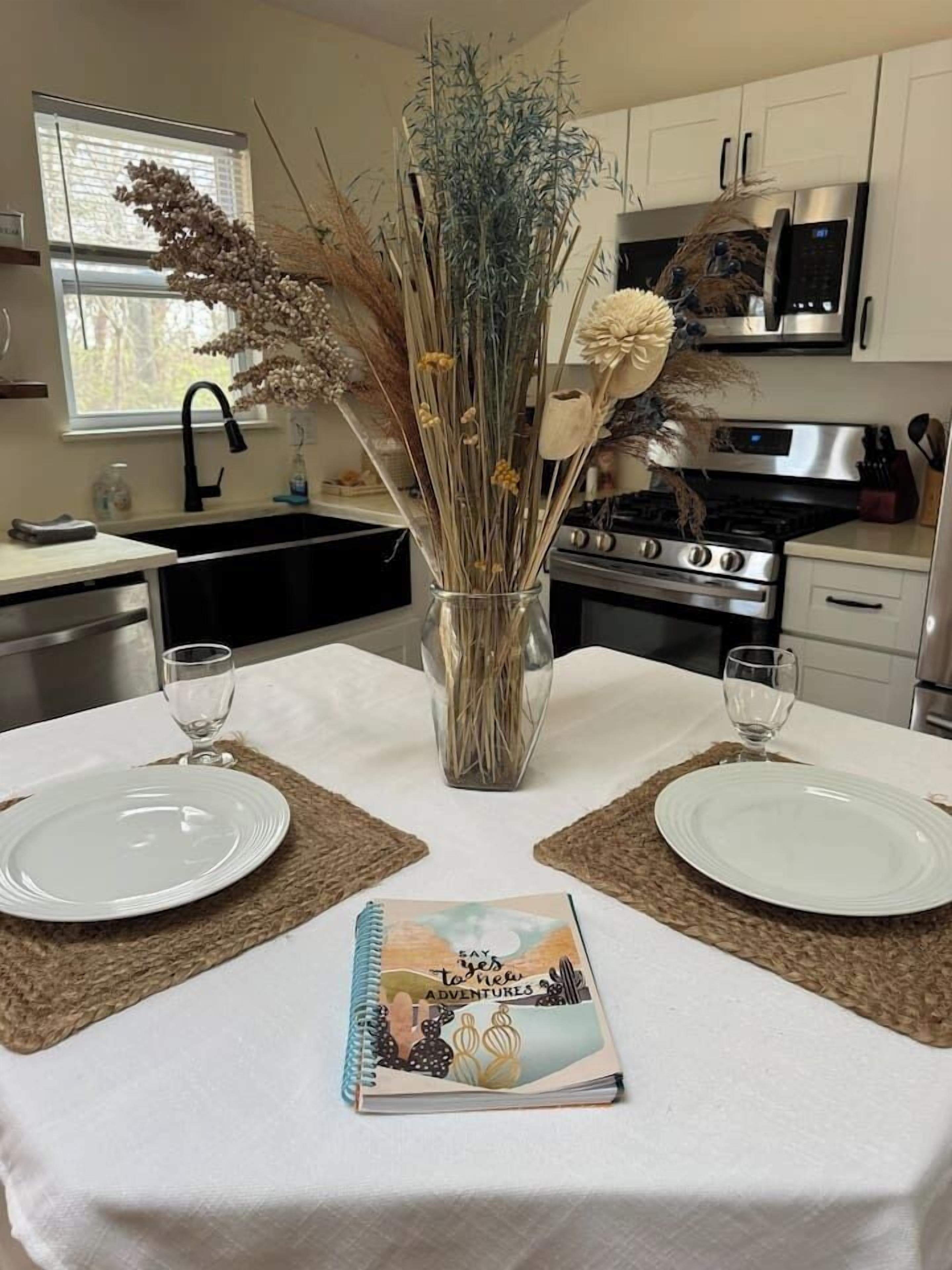 A dining table set with two plates, a vase with dried flowers, and a notebook.