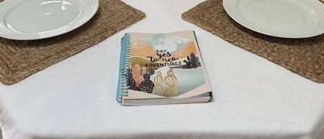 A dining table set with two plates, a vase with dried flowers, and a notebook.