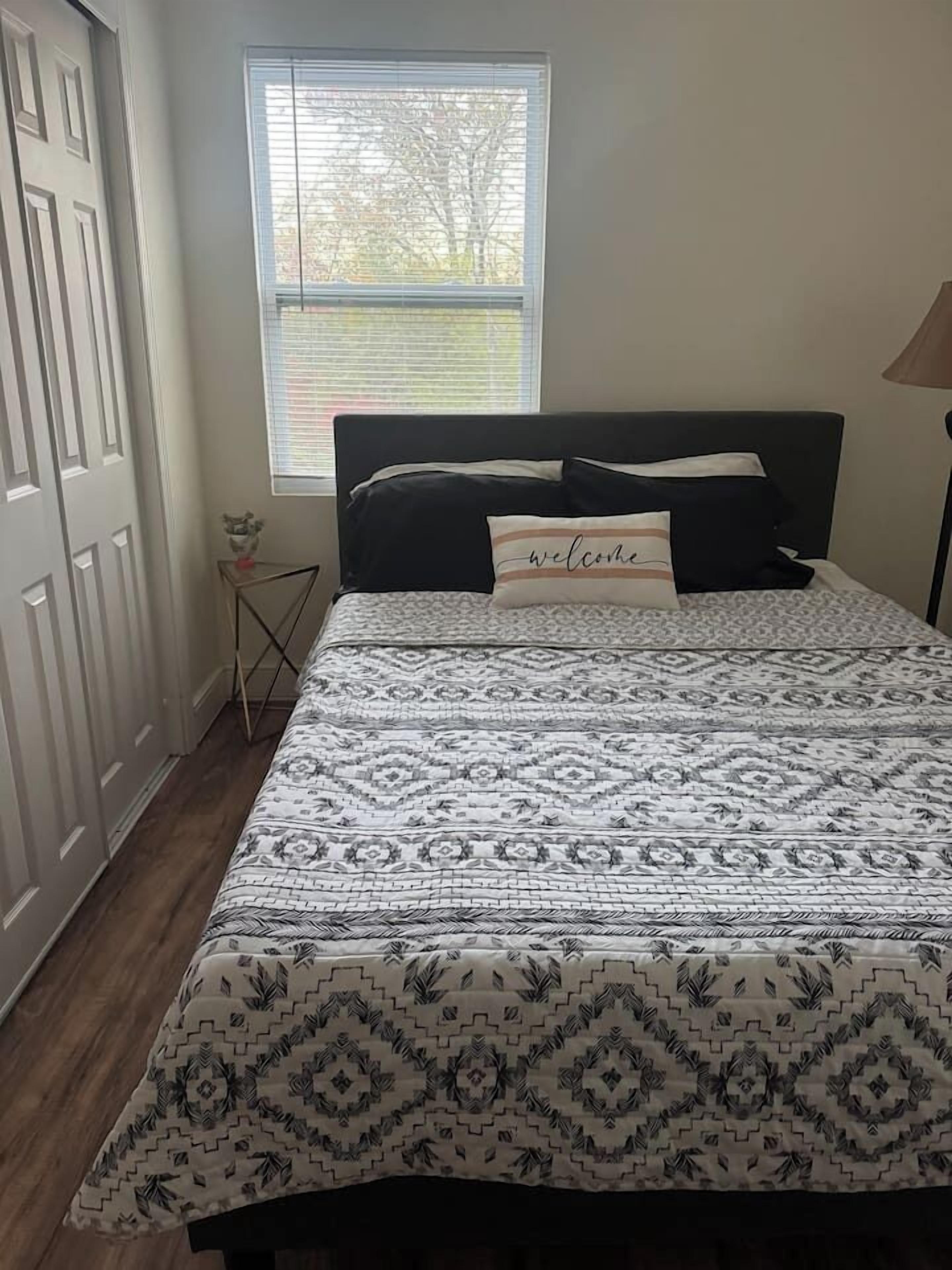 A bedroom with a bed, a window, a door, and a small table with a vase.