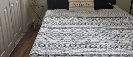 Un dormitorio con cama, ventana, puerta y una mesita de noche con un florero.