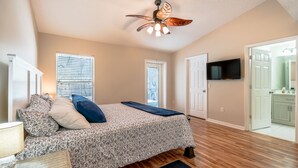 Une chambre à coucher comprenant un lit, un ventilateur de plafond, un téléviseur fixé au mur et une porte donnant sur une salle de bain.