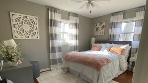 Un dormitorio con cama, escritorio, silla, ventilador de techo y ventana con cortinas.