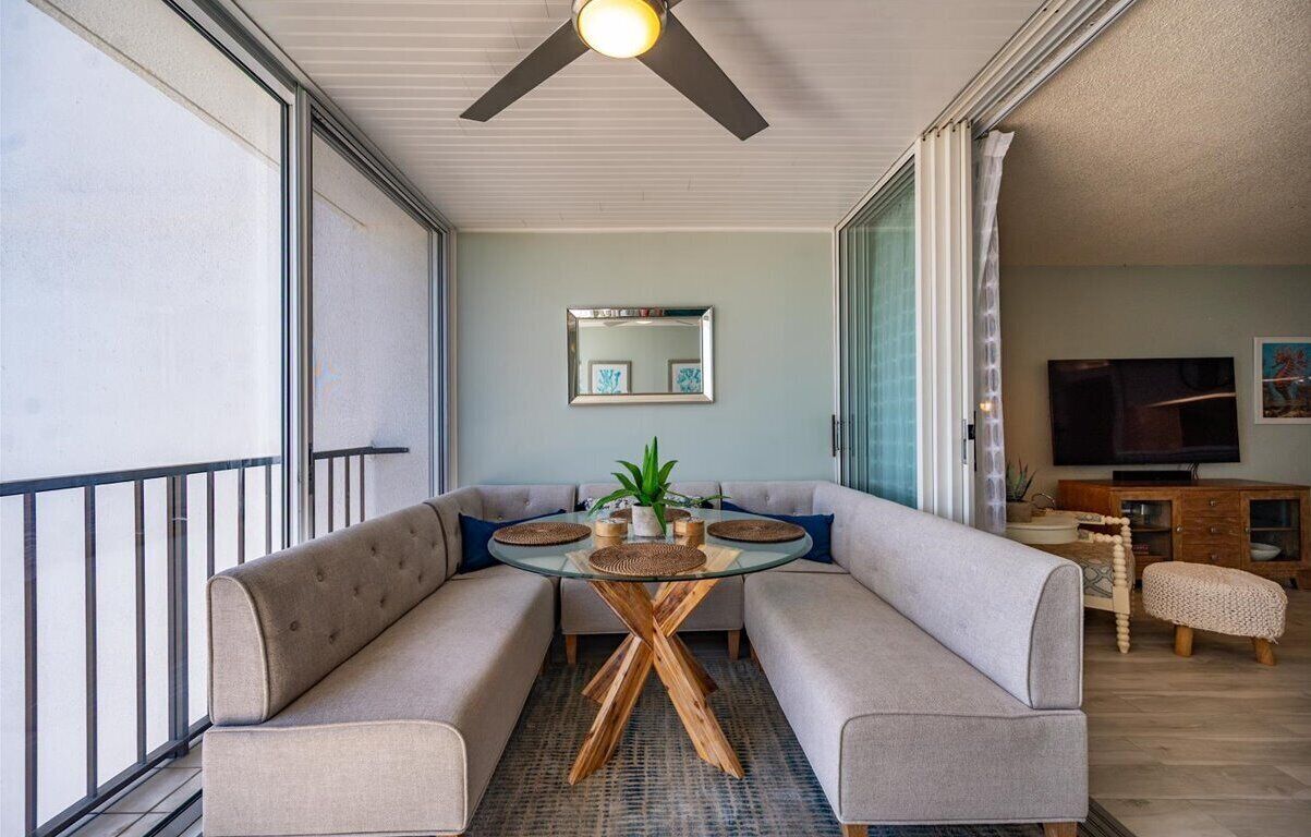 A modern living room with a glass-top coffee table, beige sofas, a ceiling fan, a flat-screen TV, and a wooden cabinet.