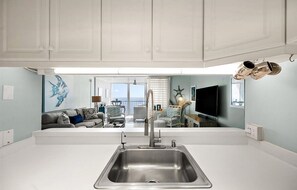 Una cocina moderna con un lavamanos grande de acero inoxidable, armarios blancos y vistas a una sala de estar con un sofá y objetos decorativos.