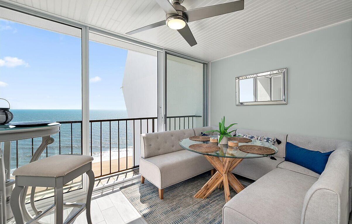 A balcony with a view of the ocean, a glass table, a sofa, and a ceiling fan.