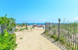 Un sendero arenoso que lleva a una zona de playa con sombrillas y gente.