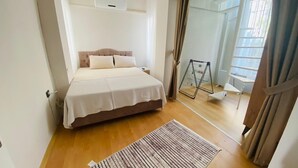 A bedroom with a bed, a bedside table, a radiator, a window with curtains, and a wooden floor. - Beautiful 3-bedroom House With Sea View and Garden (Kusadasi)