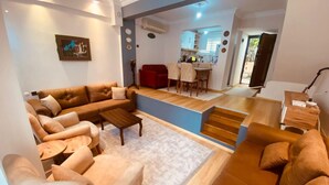 A modern living room with a sofa set, a coffee table, and a rug. There is a kitchen area visible in the background with a dining table and chairs. - Beautiful 3-bedroom House With Sea View and Garden (Kusadasi)