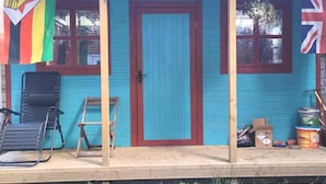 Un edificio pequeño de una sola planta con fachada azul y puerta roja. Hay una silla negra y una escalera de madera en el porche.