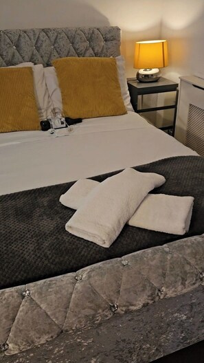 A neatly made bed with a grey headboard, a bedside lamp, and a small table with a lamp. - 2-bed Apartment, Sleeps 4,on-site Parking (Manchester)