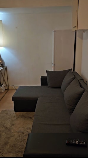 A modern living room with a sectional sofa, a small side table, a lamp, and a remote control on the sofa. - 2-bed Apartment, Sleeps 4,on-site Parking (Manchester)